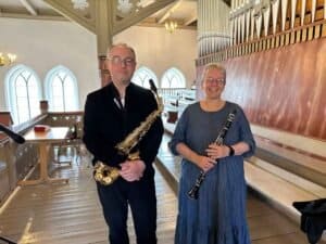 En mann som holder en saksofon og en kvinne som holder en klarinett står side om side i en kirke med trebenker og et orgel i bakgrunnen.