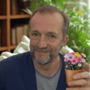 En eldre mann sitter i en sofa og holder en liten potteplante med rosa blomster. Bokhyller og grønne planter er synlige i bakgrunnen.
