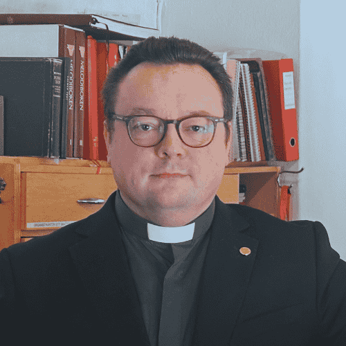Prest i svart clerical med brunt hår og briller foran bokhylle med kirkelige bøker.