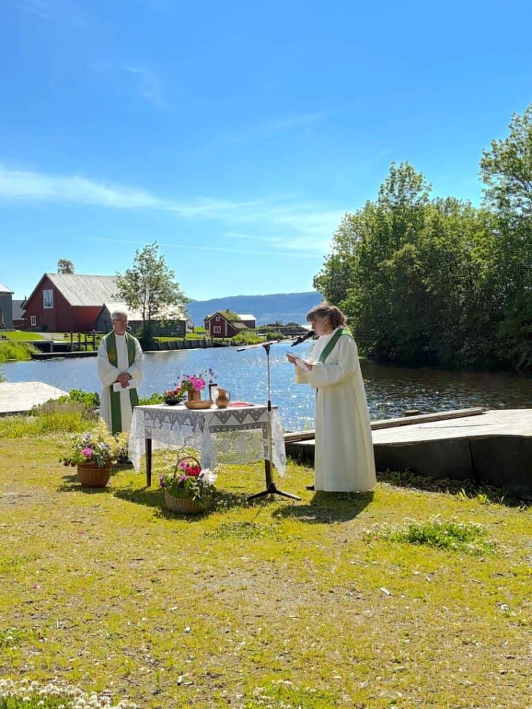 Kirkekonsert i Fosen med prest og sanger ved vannet, omgitt av natur og tradisjonelle bygninger.